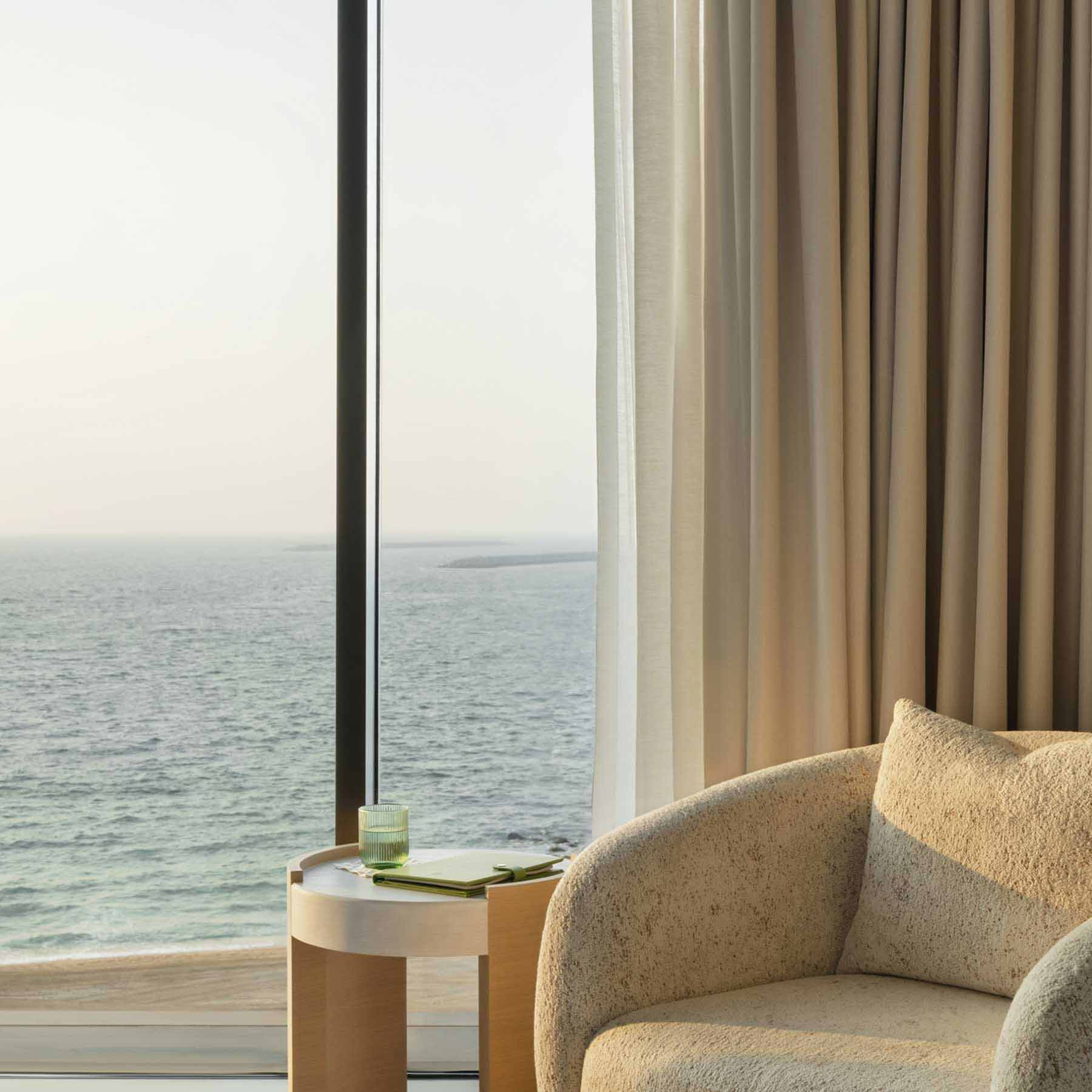Cozy seating nook by a floor-to-ceiling window with a stunning ocean view. A plush cream-colored armchair is paired with a round wooden side table holding a drink and a book. Soft beige curtains frame the window, creating a calm and relaxing atmosphere.