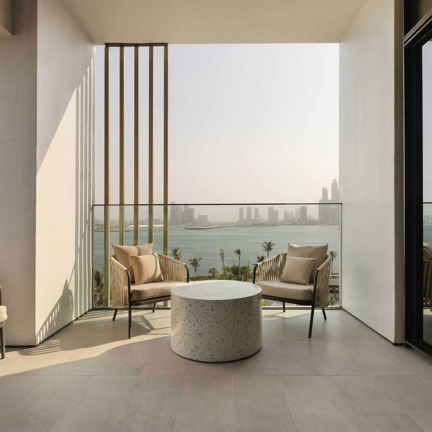 Spacious balcony with two modern armchairs and a round terrazzo coffee table, overlooking the ocean and a city skyline with high-rise buildings. Vertical slats partially frame the view, while palm trees and water below enhance the serene, resort-like atmosphere.