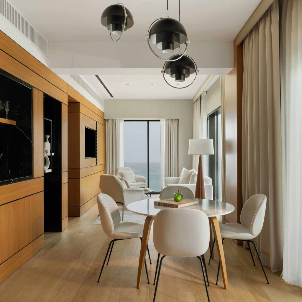 Contemporary dining area in a hotel suite, featuring a round wooden table surrounded by four white chairs with black legs. Pendant lights hang from the ceiling, while a floor lamp and abstract artwork decorate a wood-paneled accent wall. Floor-to-ceiling curtains and built-in cabinetry enhance the modern, warm ambiance.