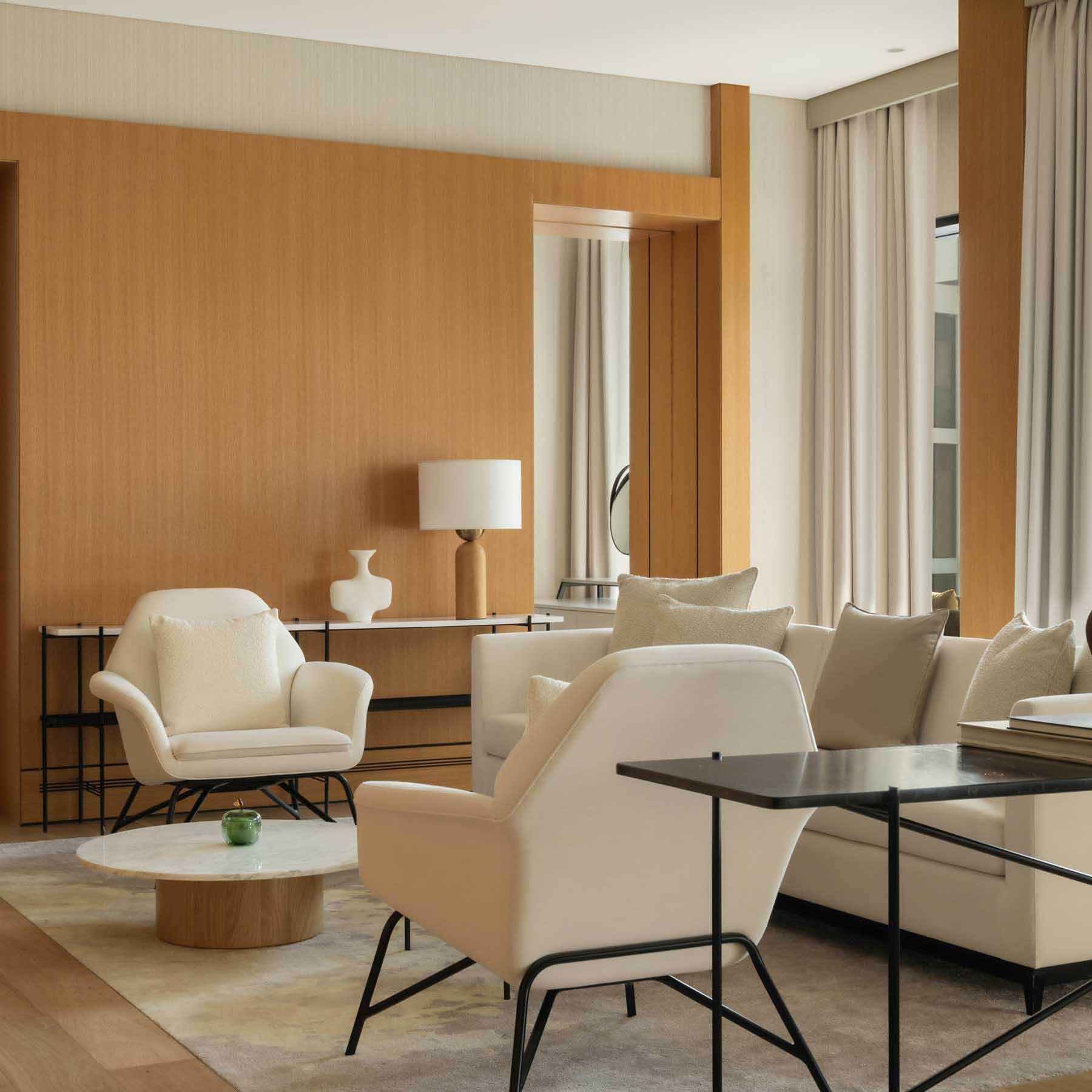 Stylish hotel suite featuring a lounge area with two white armchairs, a black side table with books, a small round coffee table, and a built-in wooden entertainment unit with a television. A hallway leads to a bedroom with a modern design, while warm wood tones and neutral decor create a sophisticated ambiance.