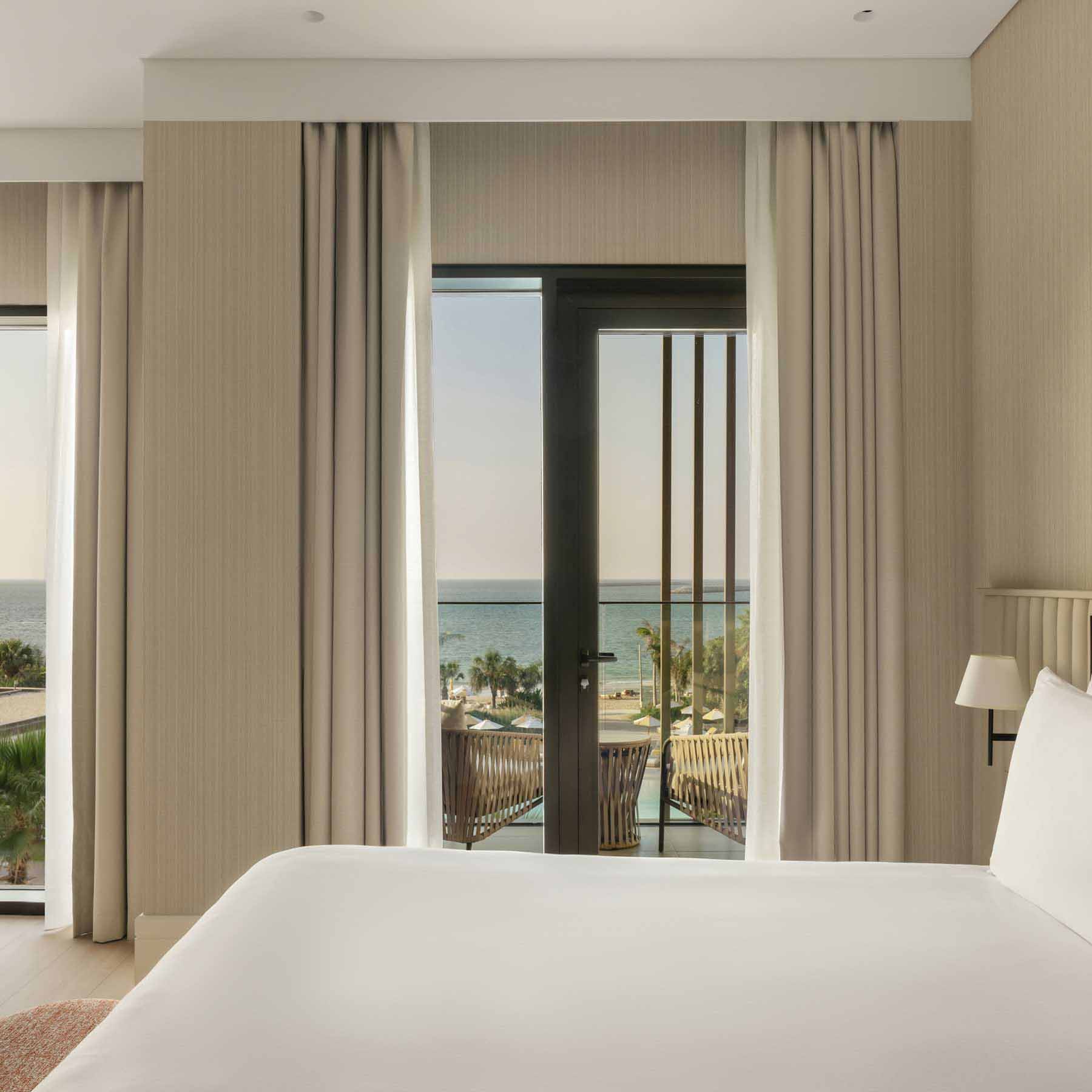 Modern hotel room with a king-size bed, a vanity desk with a round mirror, and floor-to-ceiling windows offering a view of the ocean and palm trees. A balcony with two chairs and a table overlooks the sea, while soft curtains and wooden accents create a calming, elegant atmosphere.