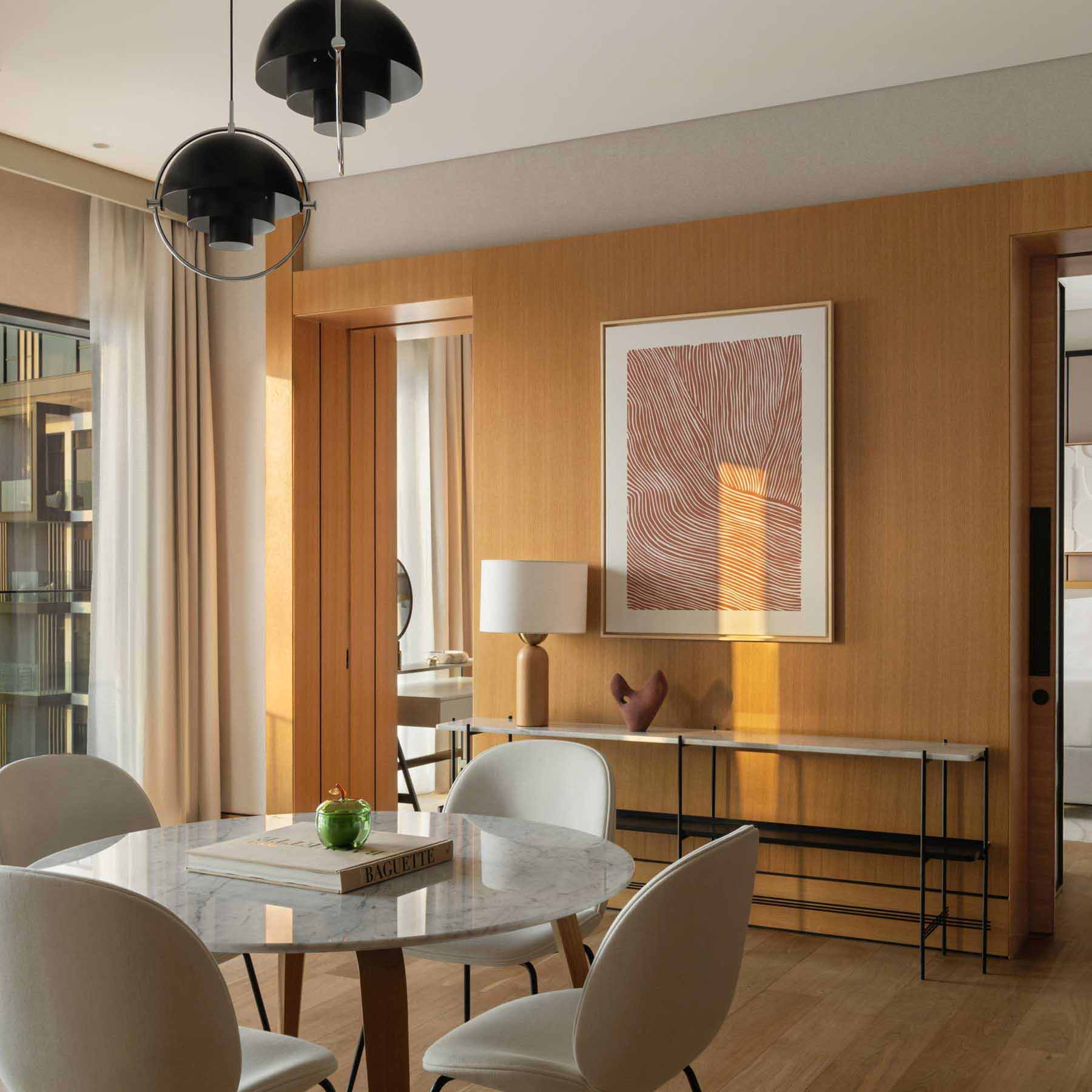 Contemporary hotel suite with an open-plan living and dining area, featuring a round marble dining table with four white chairs, a beige sofa with cushions, a wooden coffee table, and a floor lamp. Large windows with curtains let in natural light, and a mirror wall reflects the modern interior design.