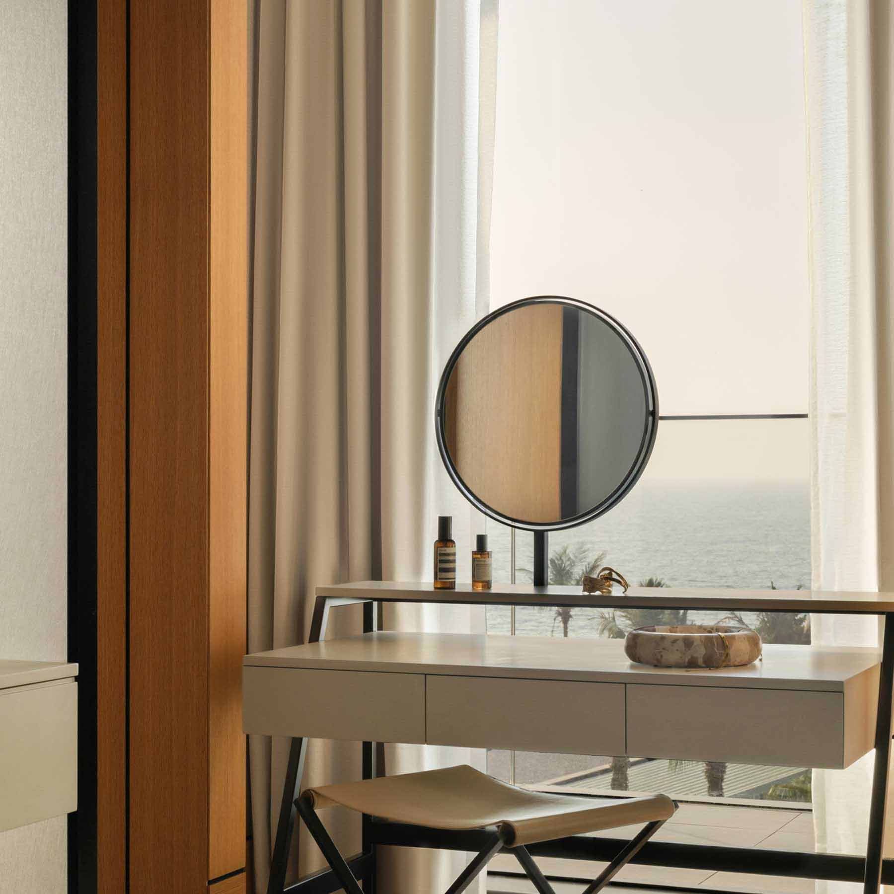 a vanity with a round mirror facing the window with views of the sea
