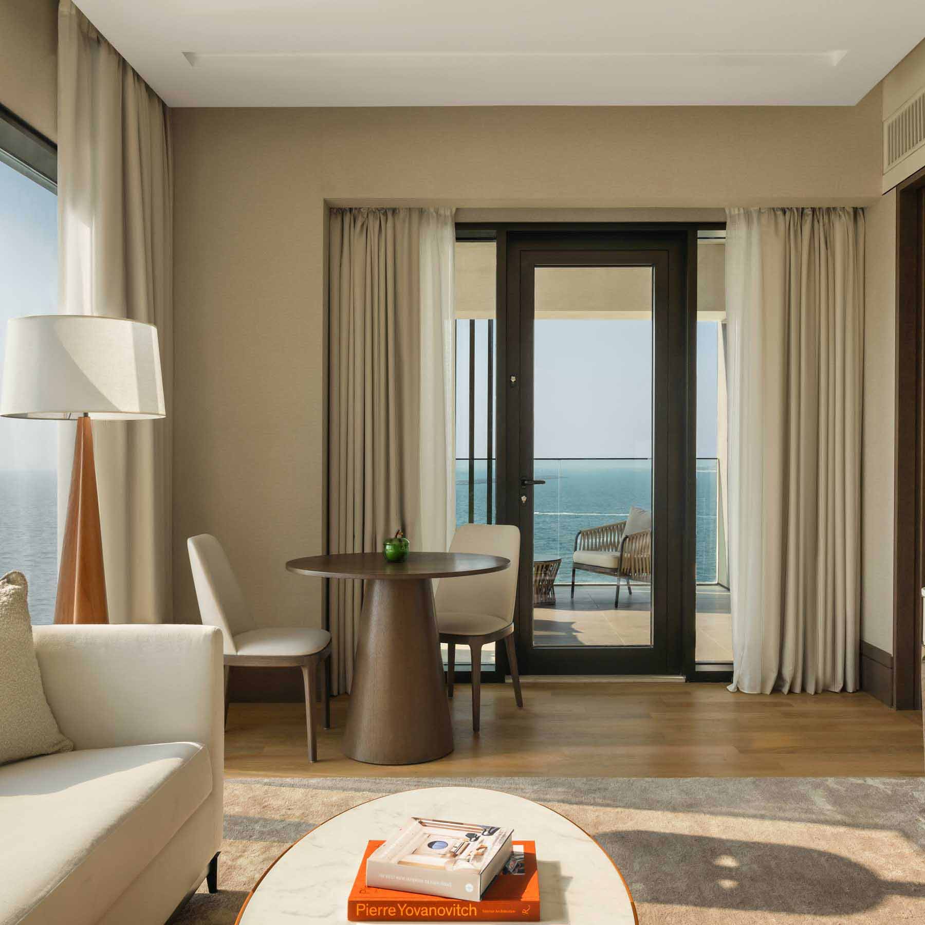 a guestroom with a white couch, coffee table and dining table in the corner of the room with two chairs and sliding glass doors that lead to a balcony with sea views