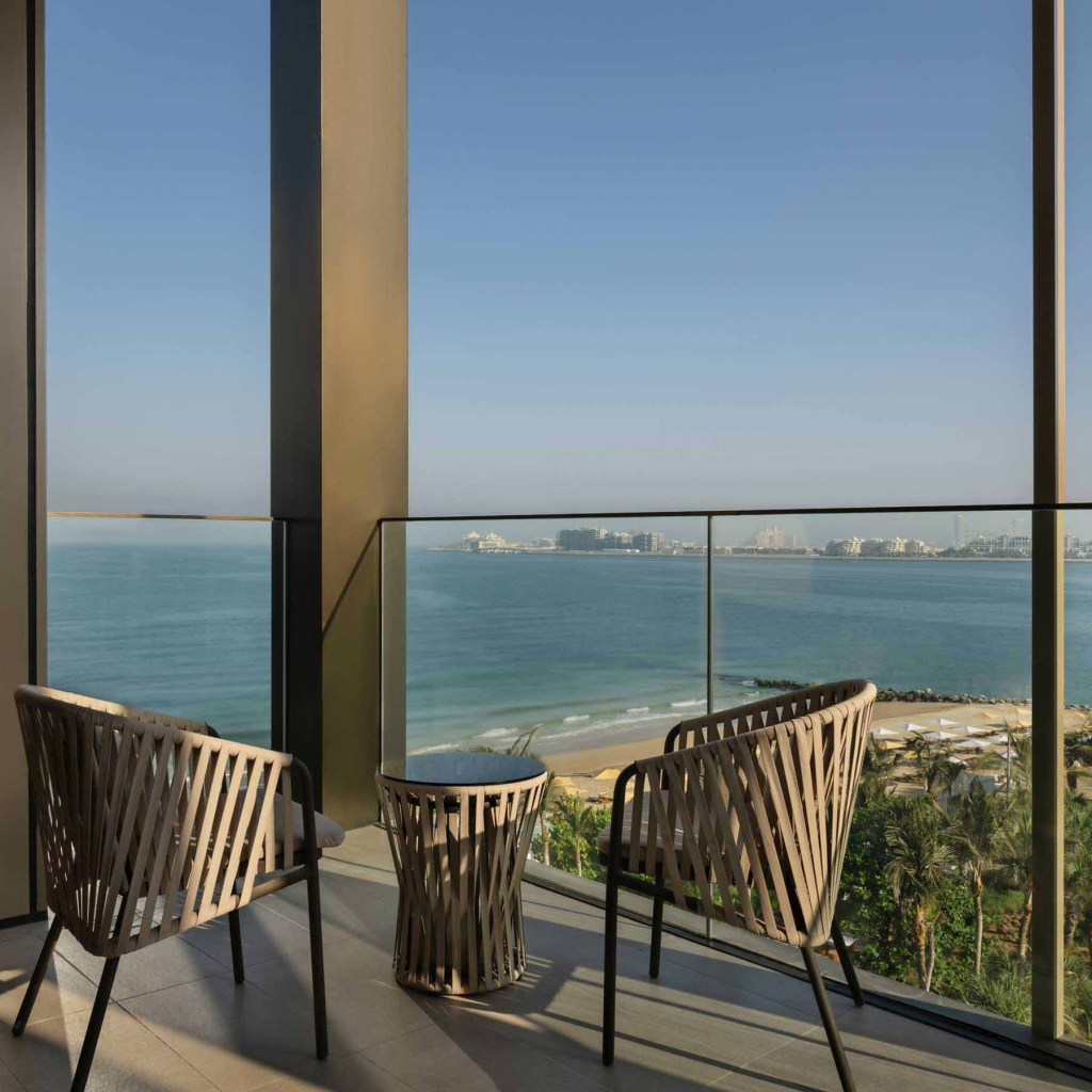 Two wicker chairs and a table rest on a balcony, overlooking a tranquil beach and sea, framed by palm trees at the Delano Hotel, Dubai.