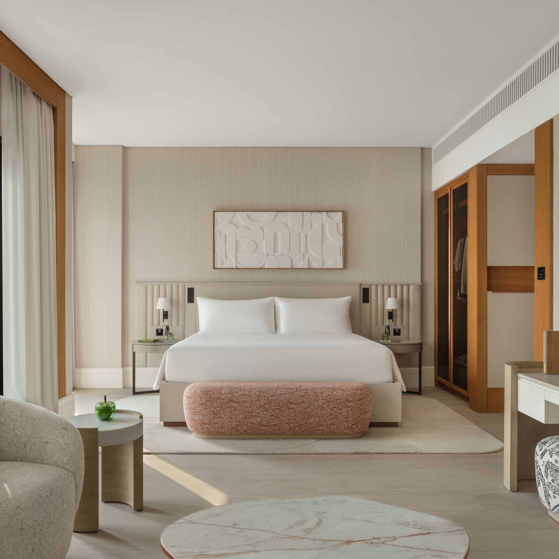 Elegant hotel bedroom featuring a king-size bed with crisp white linens, a textured pink bench at the foot, and modern bedside tables with matching lamps. A built-in open wardrobe with wooden accents is visible, alongside a seating area with a round marble coffee table. Neutral tones, soft textures, and natural light create a calming, sophisticated atmosphere.