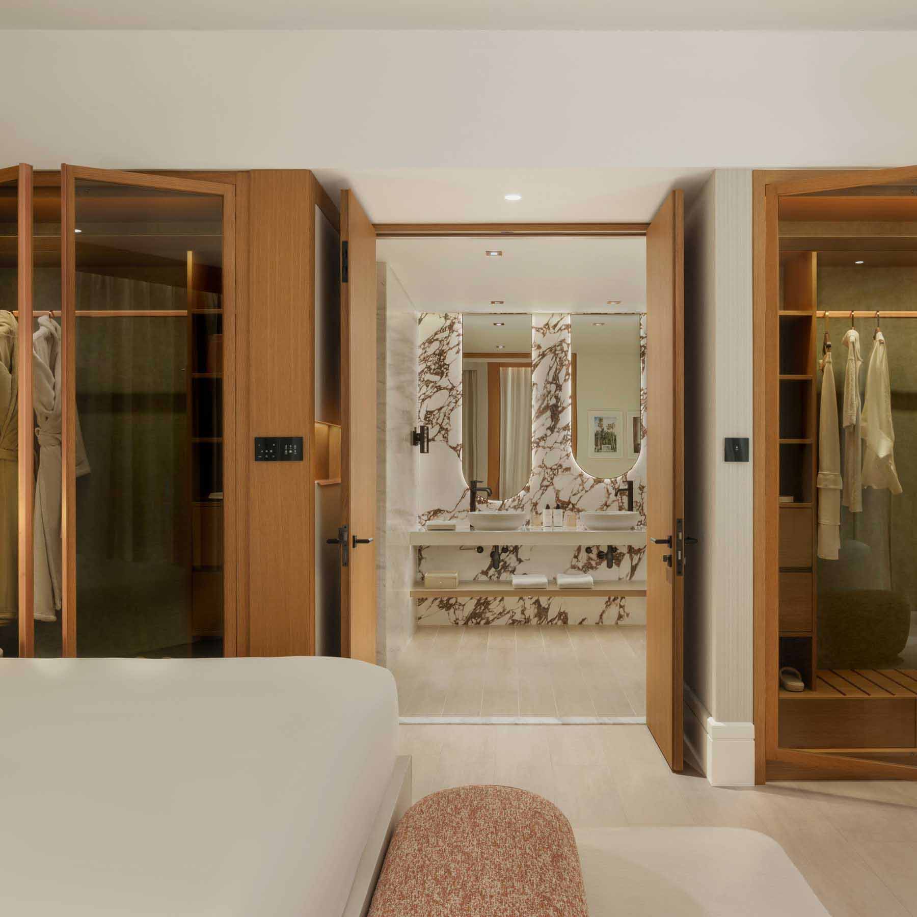 Modern hotel bedroom with an open wooden wardrobe featuring glass doors, illuminated interior, and neatly arranged clothing and shoes. A vanity area with a curved stool, marble countertop, and oval mirror is visible in the background. Neutral tones and wooden accents create a warm, sophisticated ambiance.