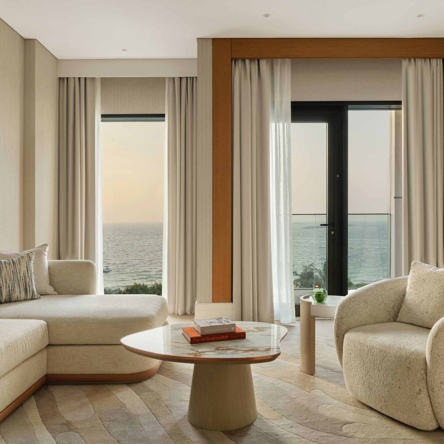 un espace de vie luxueux avec un canapé blanc sectionnel, une table basse en marbre et une chaise blanche, avec beaucoup de lumière naturelle provenant de la porte coulissante en verre qui mène au balcon avec vue sur la mer.