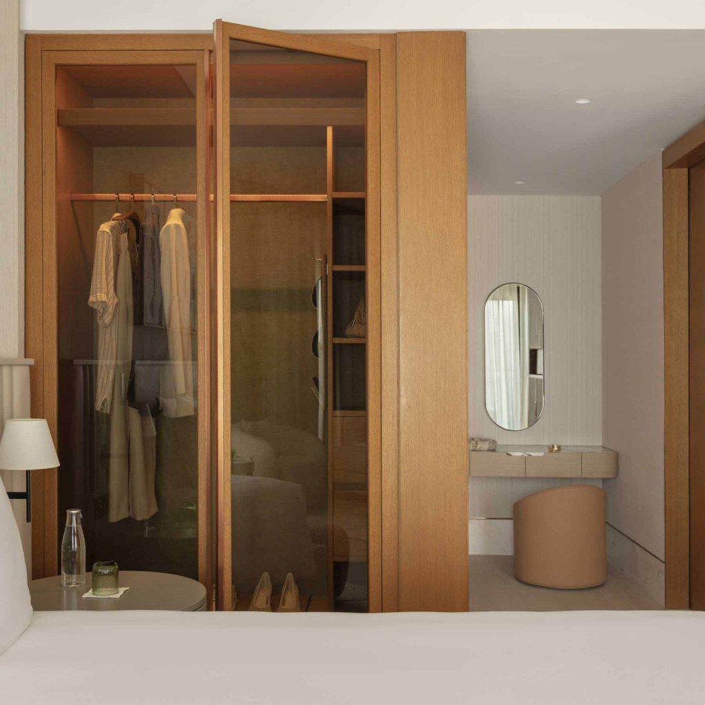 Modern hotel bedroom with an open wooden wardrobe featuring glass doors, illuminated interior, and neatly arranged clothing and shoes. A vanity area with a curved stool, marble countertop, and oval mirror is visible in the background. Neutral tones and wooden accents create a warm, sophisticated ambiance.