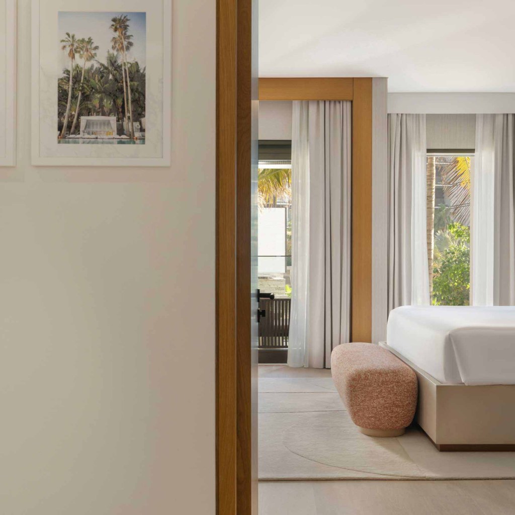 the view of the bedroom from the bathroom with the foot of the bed visible and the salmon colored bench