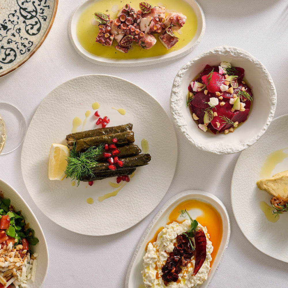 Assorted Mediterranean mezze dishes elegantly arranged on a white tablecloth, including stuffed grape leaves garnished with pomegranate, marinated octopus, beetroot salad with nuts and herbs, creamy cheese dip with chili oil, and other vibrant appetisers served in textured ceramic plates.