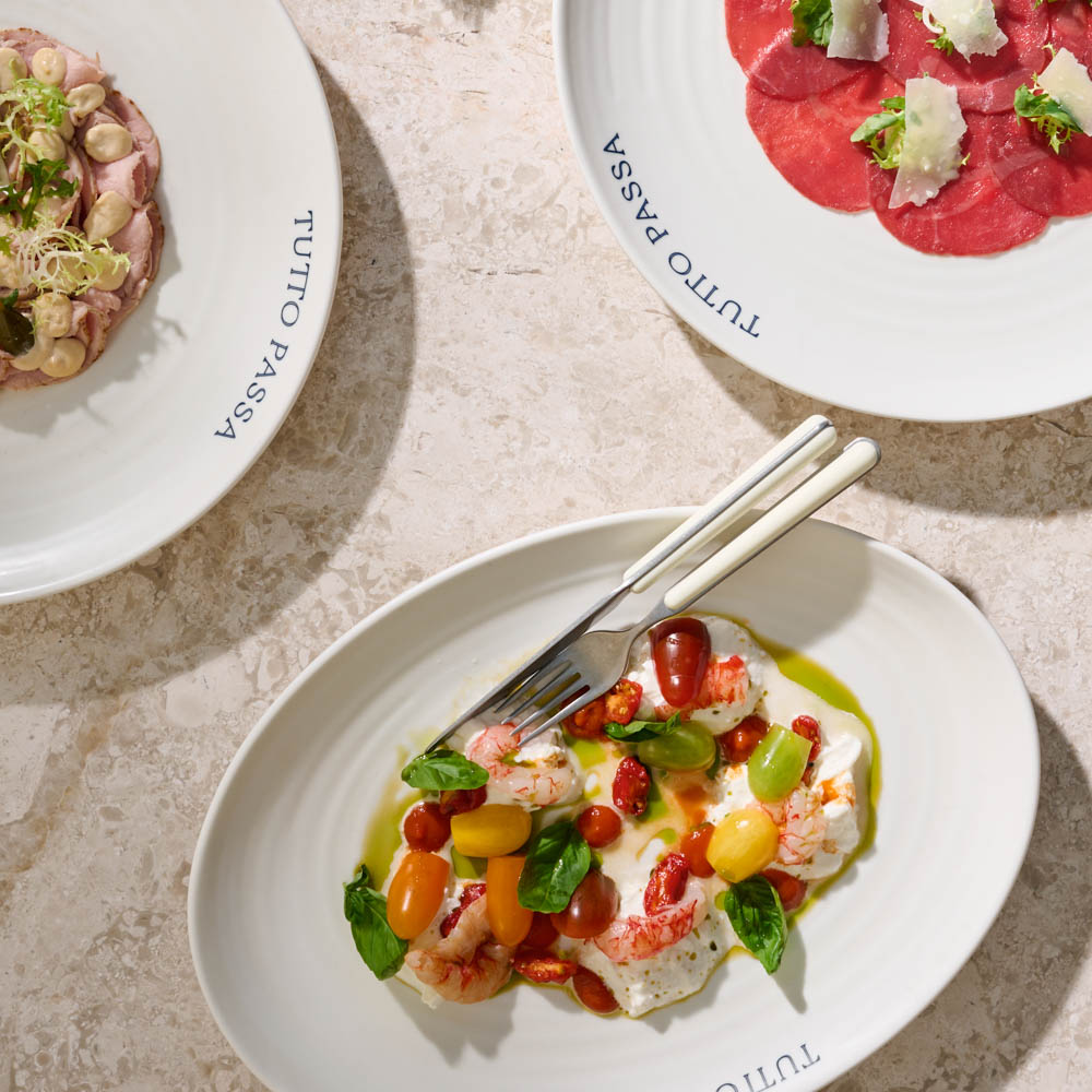 Flat lay of three elegantly plated Italian appetisers on white ceramic dishes, including beef carpaccio with parmesan, tuna with creamy sauce and capers, and burrata with cherry tomatoes and basil, served on a stone countertop. Plates are branded with 'TUTTO PASSA'.