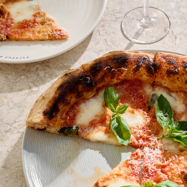 Close-up of a freshly baked Neapolitan-style Margherita pizza topped with melted mozzarella, tomato sauce, grated parmesan, and fresh basil leaves. A slice is served on a side plate beside a glass of white wine, all placed on a light marble table.