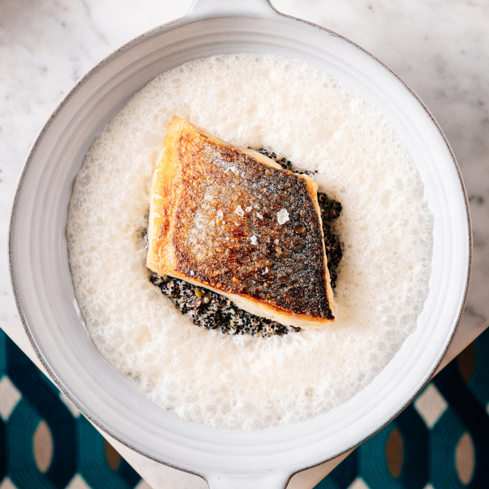 Top-down view of a gourmet dish featuring a perfectly seared fillet of fish with crispy skin, placed atop a bed of black quinoa and surrounded by a delicate white foam sauce, served in an elegant ceramic bowl on a marble surface.