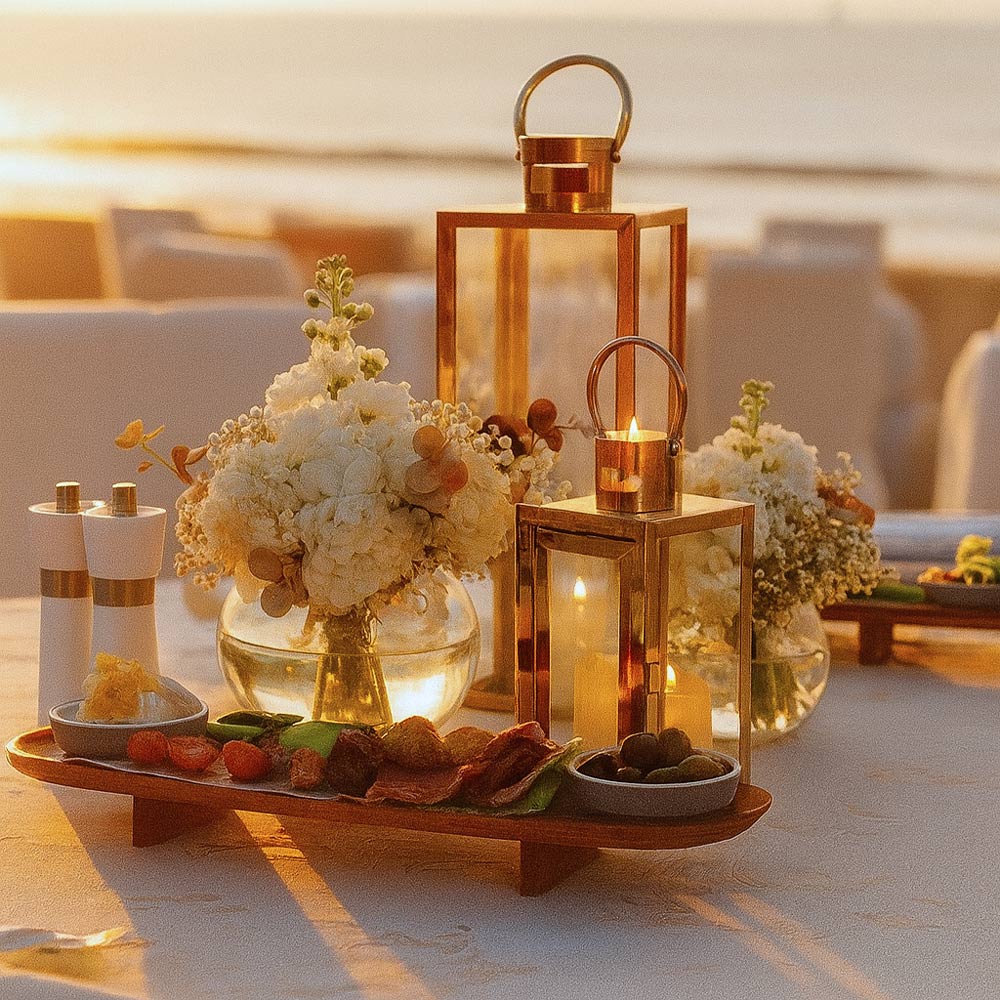 Elegant beachside table setup at sunset featuring gold lanterns with lit candles, fresh white floral arrangements, and a refined selection of appetisers on a wooden platter.