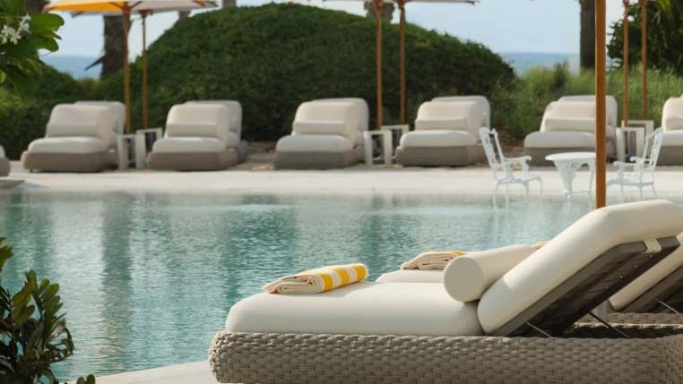 Luxury poolside setting with plush lounge chairs, yellow and white sun umbrellas, and swaying palm trees, overlooking a serene beachside landscape, creating a tropical and relaxing atmosphere.
