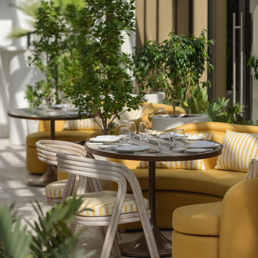 Elegant outdoor restaurant seating with round tables, yellow cushioned sofas, striped pillows, and greenery creating a fresh, inviting atmosphere.
