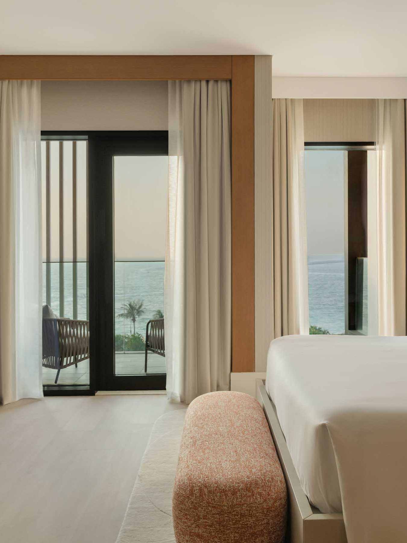 the side view of a king bed with a salmon colored bench at the foot, with views of the sea out the floor to ceiling windows.