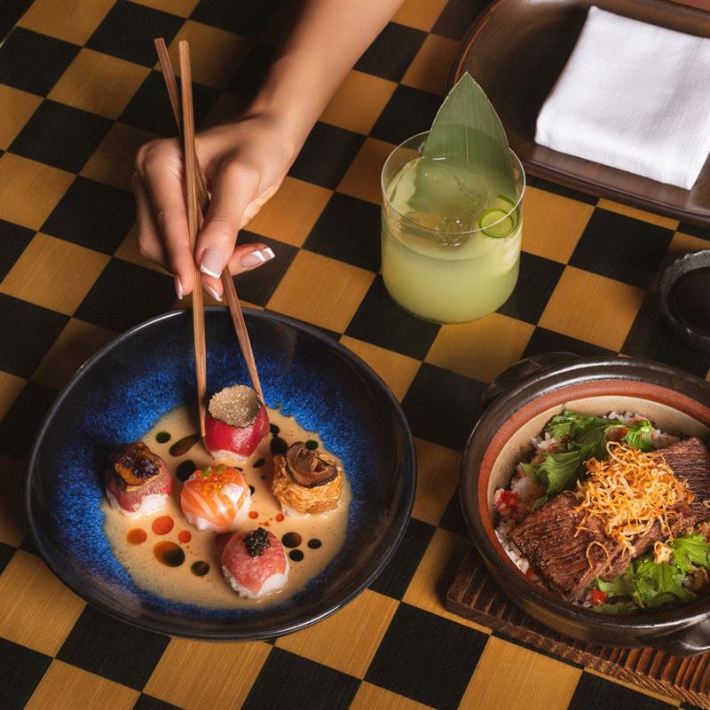 Hand using chopsticks to pick sushi from an elegant bowl, accompanied by a green cocktail and a Japanese rice dish on a checkered table setting.