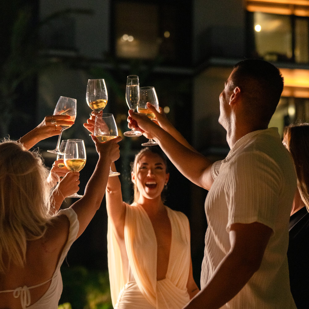 Group of elegantly dressed friends raising glasses in a celebratory toast during an evening outdoor event, illuminated by warm lights.