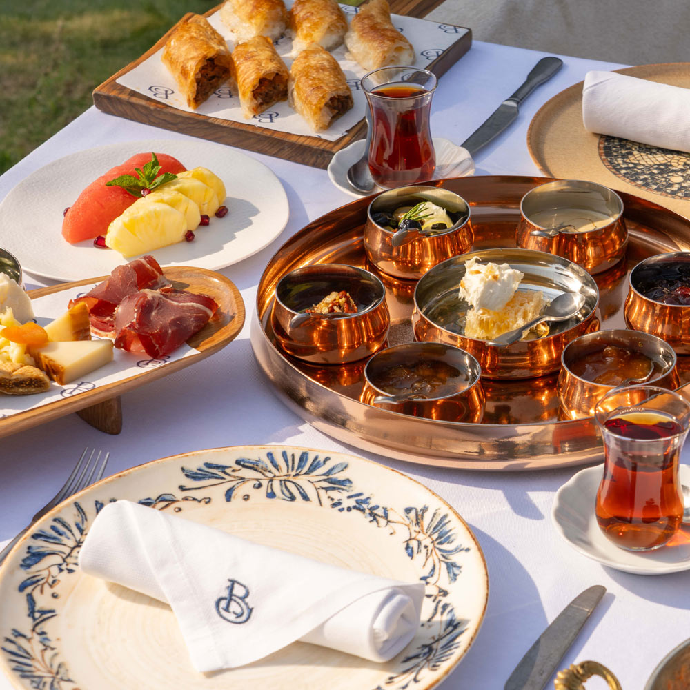 Traditional Middle Eastern breakfast spread served outdoors, featuring assorted cheeses, cured meats, honeycomb, pastries, fresh fruit, and Turkish tea in glass cups on a white table setting.