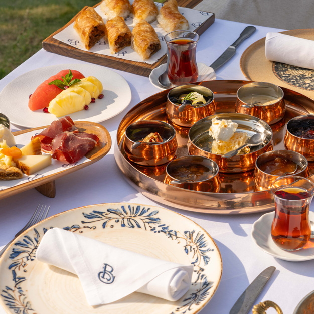 Traditional Middle Eastern breakfast spread served outdoors, featuring assorted cheeses, cured meats, honeycomb, pastries, fresh fruit, and Turkish tea in glass cups on a white table setting.