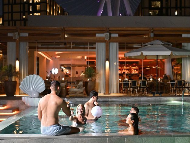 Group of friends enjoying a lively evening swim in a lit-up pool at a luxury venue in Dubai, with the illuminated Ain Dubai Ferris wheel glows in the background, and the chic indoor bar and dining space