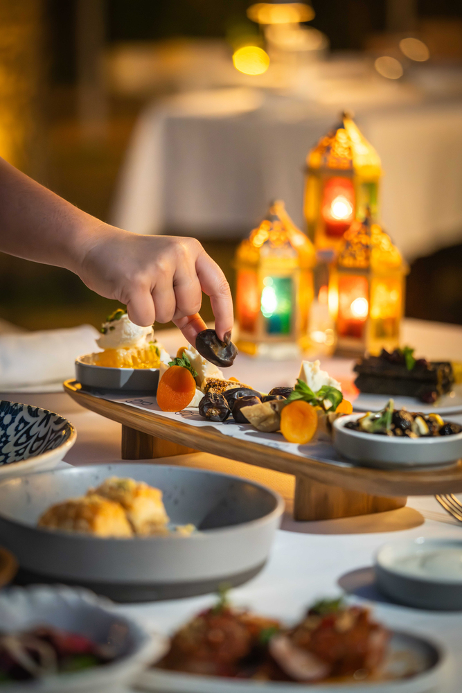 A guest selecting dates from an elegant Middle Eastern iftar spread, with traditional lanterns glowing in the background and a beautifully presented assortment of mezze and desserts on the table.