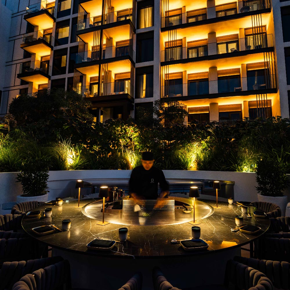 Outdoor teppanyaki dining experience at Delano Dubai, featuring a circular chef’s table set in a landscaped courtyard, with a live teppanyaki chef at the grill and illuminated hotel balconies in the background at night.