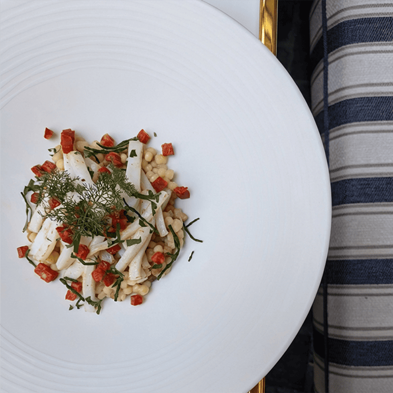 White plate with fregola sarda, squid, chorizo. Tha plate sits on a marble table with a golden frame, on the right side you can see a blue and white striped cushion, at Maison Delano Paris.