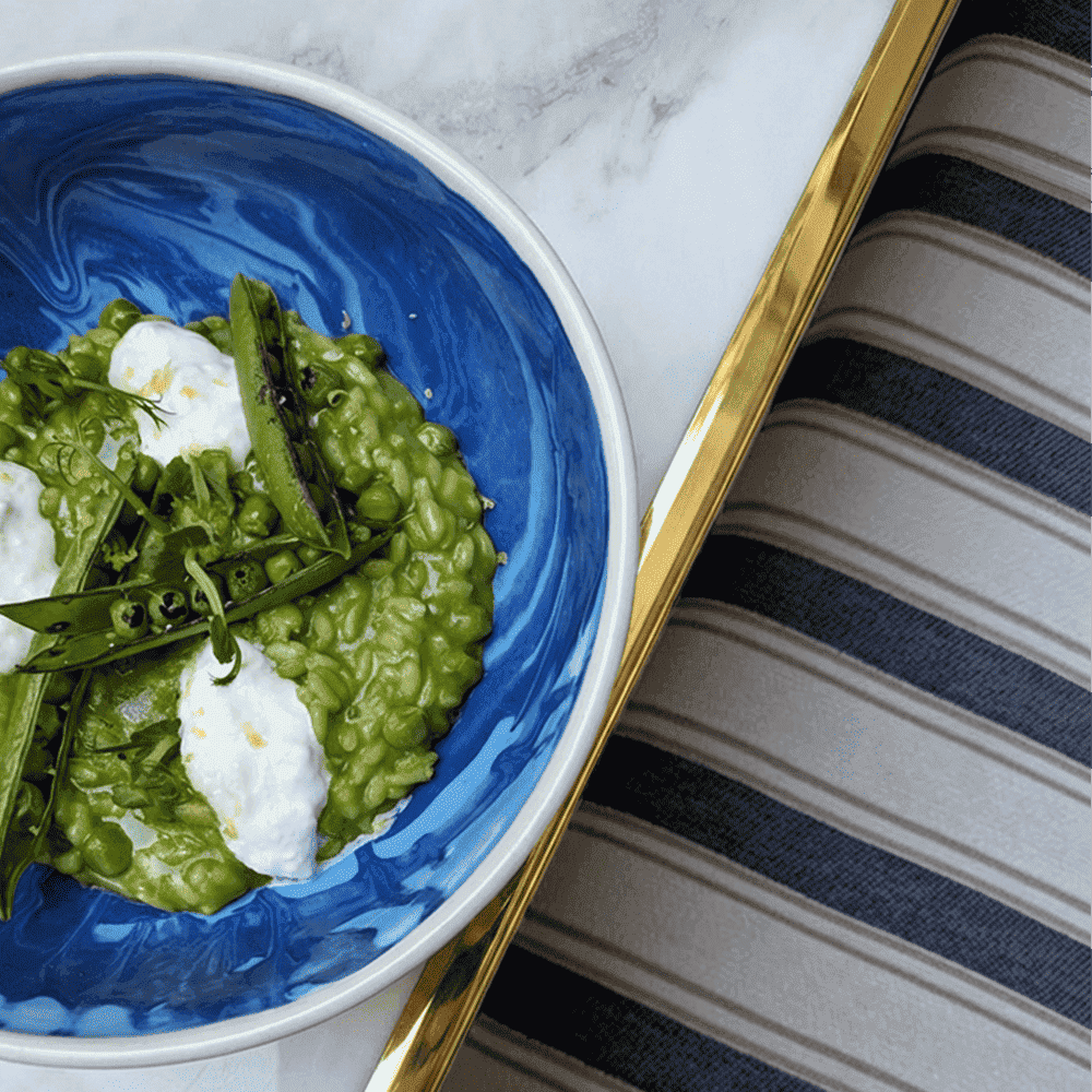 On a marble table, with a golden frame, we can see a blue plate with a green risotto. In top, we can see some green peas in the peals and some straciatella. On the right side of the pictire, we can see a blue and white striped cushion, at Maison Delano Paris.